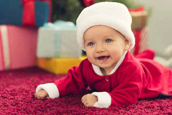 Laughing little baby in santa suit lying on red carpet with christmas gifts blurred on background — Stock Photo