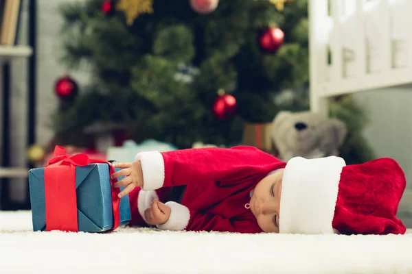 Adorable petit bébé en costume de Père Noël couché sur le sol avec boîte cadeau de Noël — Photo de stock