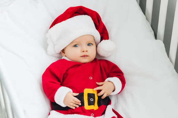 Blick aus der Vogelperspektive auf ein entzückendes kleines Baby im Weihnachtsmann-Anzug, das im Bett liegt und in die Kamera schaut — Stockfoto