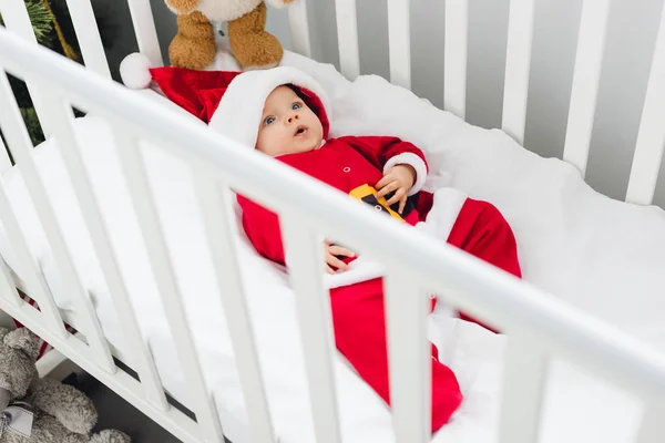 Vue grand angle de mignon petit bébé en costume de Père Noël couché dans la crèche avec ours en peluche et regardant la caméra — Photo de stock