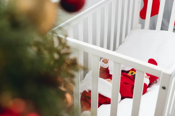 Entzückendes kleines Baby im Weihnachtsmannanzug in der Krippe liegend mit Weihnachtsbaum im Vordergrund — Stockfoto