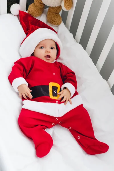 Blick aus der Vogelperspektive auf ein entzückendes kleines Baby im Weihnachtsmannanzug, das mit Teddybär in der Krippe liegt und in die Kamera schaut — Stockfoto