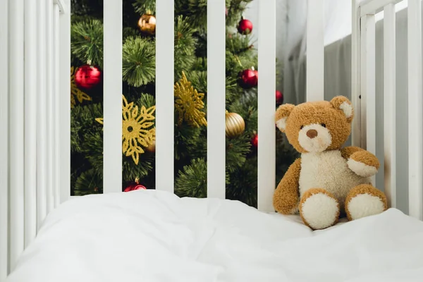 Ursinho de pelúcia na cama infantil na frente da árvore de natal — Fotografia de Stock