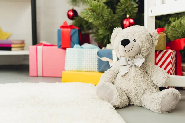 Primer plano de osito de peluche apoyado en la cuna delante del árbol de Navidad y regalos - foto de stock