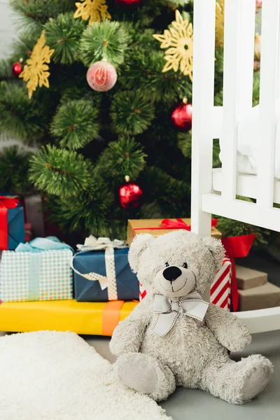 Ursinho de pelúcia apoiando-se no berço na frente da árvore de natal e presentes — Fotografia de Stock