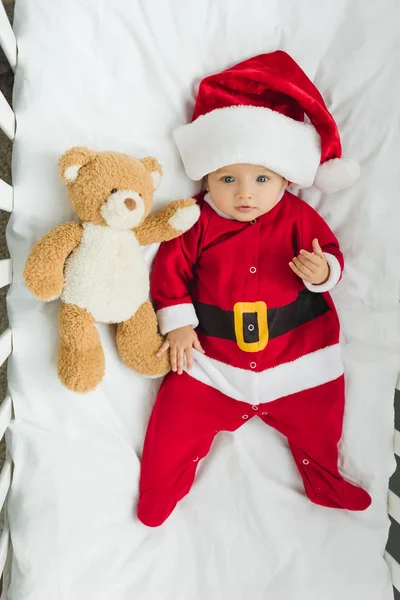 Blick von oben auf das süße kleine Baby im Weihnachtsmannanzug, das mit Teddybär in der Krippe liegt und in die Kamera schaut — Stockfoto