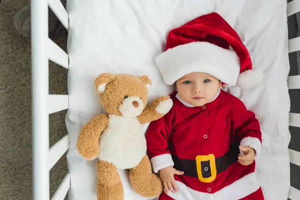Vue de dessus du beau petit bébé en costume de Père Noël couché dans la crèche avec ours en peluche et regardant la caméra — Photo de stock