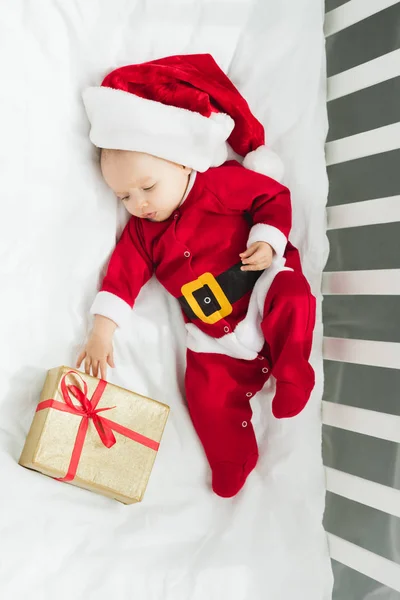 Top view of beautiful little baby in santa suit lying in crib with christmas gift — Stock Photo