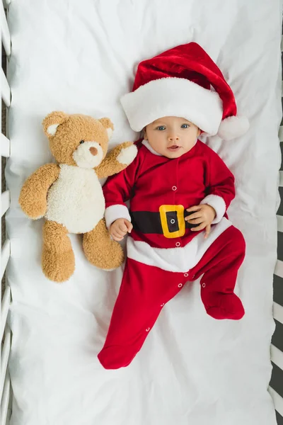 Top view of beautiful little baby in santa suit lying in crib with teddy bear — Stock Photo