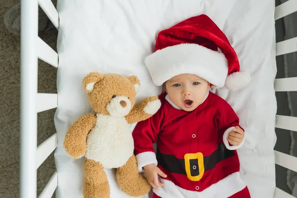 Top view of happy little baby in santa suit lying in crib with teddy bear — Stock Photo