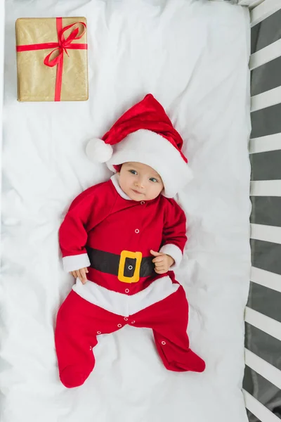 Top view of happy little baby in santa suit lying in crib with christmas gift — Stock Photo