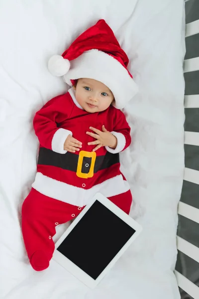 Top view of beautiful little baby in santa suit lying in crib with tablet — Stock Photo