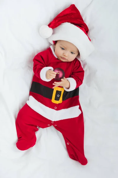 Top view of beautiful little baby in santa suit holding christmas bauble while lying in crib — Stock Photo