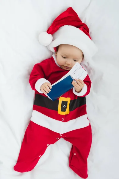 Вид на симпатичного малыша в костюме Санты лежащего в кроватке с авиабилетом и паспортом — стоковое фото