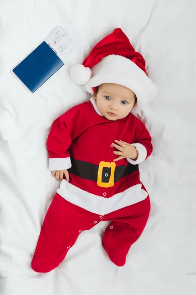 Vista dall'alto dell'adorabile bambino in costume da Babbo Natale sdraiato in culla con biglietto aereo e passaporto — Foto stock