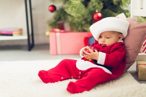 Adorable little baby in santa suit sitting on floor with blurred christmas tree on background — Stock Photo
