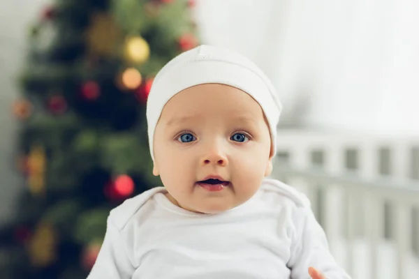 Close-up portrait of adorable little baby in white hat looking at camera with blurred christmas tree on background — Stock Photo