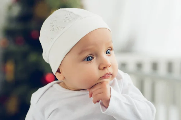 Close-up portrait of cute little baby in white hat with blurred christmas tree on background — Stock Photo