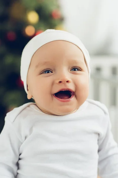 Close-up portrait of laughing little baby in white hat with blurred christmas tree on background — Stock Photo