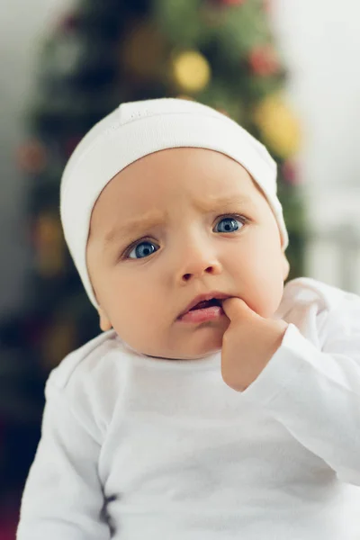 Retrato de primer plano de bebé confundido en sombrero blanco con árbol de navidad borrosa en el fondo - foto de stock