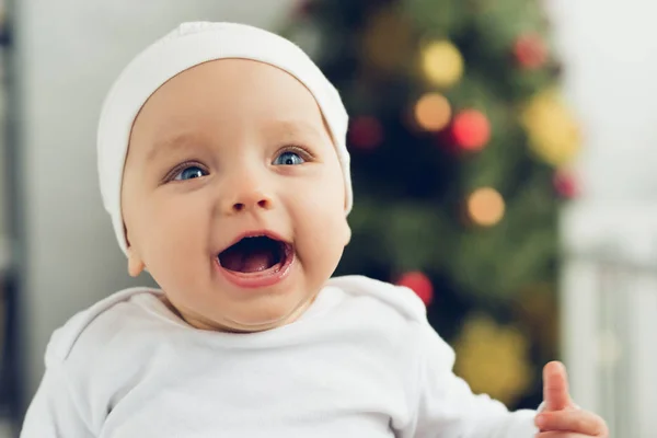 Nahaufnahme Porträt eines emotionalen kleinen Babys mit weißem Hut und unscharfem Weihnachtsbaum auf dem Hintergrund — Stockfoto