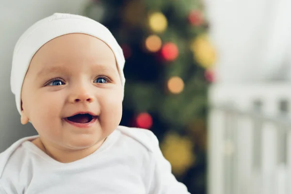 Portrait en gros plan de heureux petit bébé en chapeau blanc avec arbre de Noël flou sur le fond — Photo de stock