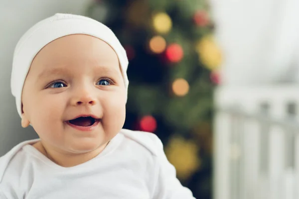 Close-up portrait of happy little baby in white hat with blurred christmas tree on background — Stock Photo