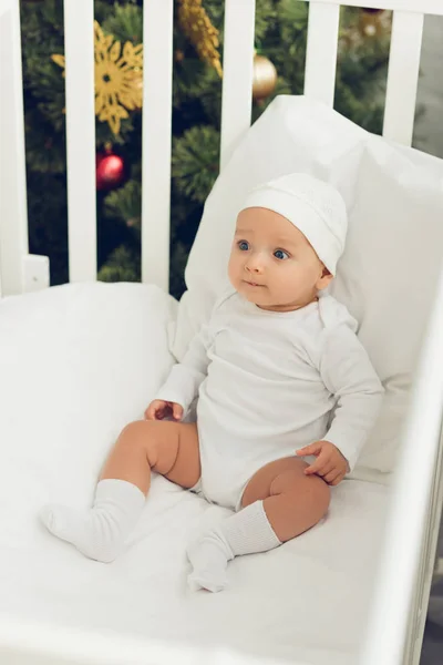 High angle view of beautiful little baby in white hat sitting in crib — Stock Photo