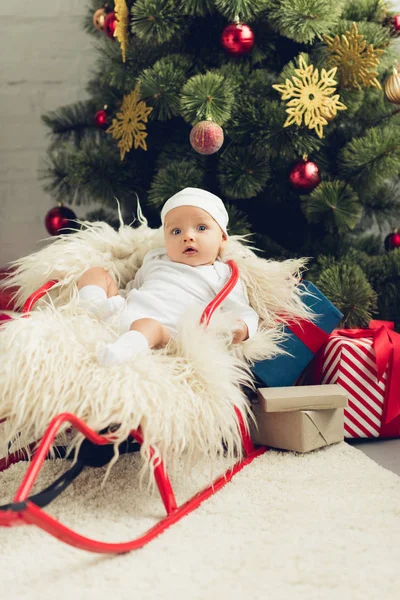 Adorable little baby in sleigh in front of christmas tree with gifts — Stock Photo