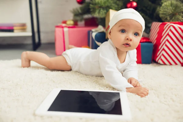 Close-up shot of beautiful little baby with tablet lying on floor with christmas gifts and tree blurred on background — Stock Photo