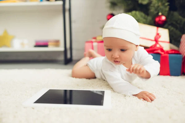 Close-up shot of cute little baby with tablet lying on floor with christmas gifts and tree blurred on background — Stock Photo