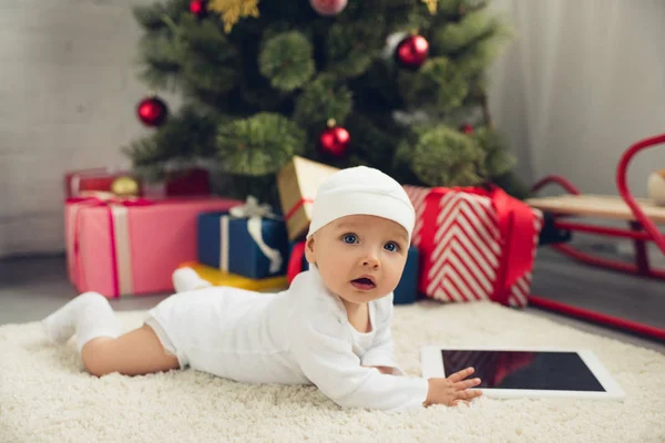 Cute little baby with tablet lying on floor with christmas gifts and tree blurred on background — Stock Photo