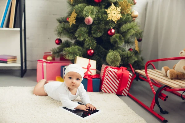 Entzückendes kleines Baby mit Tablet, das neben Weihnachtsgeschenken und Baum auf dem Boden liegt und in die Kamera schaut — Stockfoto