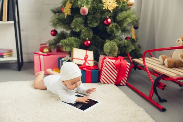 Entzückendes kleines Baby mit Tablet auf dem Boden liegend in der Nähe von Weihnachtsgeschenken und Baum — Stockfoto