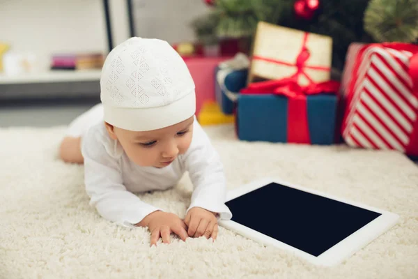 Entzückendes kleines Baby mit Tablet auf dem Boden liegend mit Weihnachtsgeschenken auf dem Hintergrund verschwommen — Stockfoto