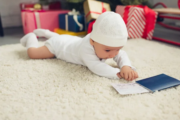 Niedliches kleines Baby mit Flugticket und Reisepass auf dem Boden liegend, Weihnachtsgeschenke im Hintergrund verschwommen — Stockfoto