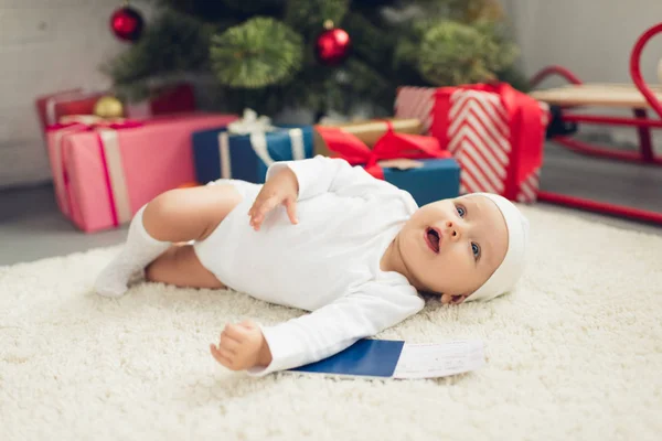 Adorable petit bébé avec billet d'avion et passeport couché sur le sol avec arbre de Noël flou sur fond — Photo de stock