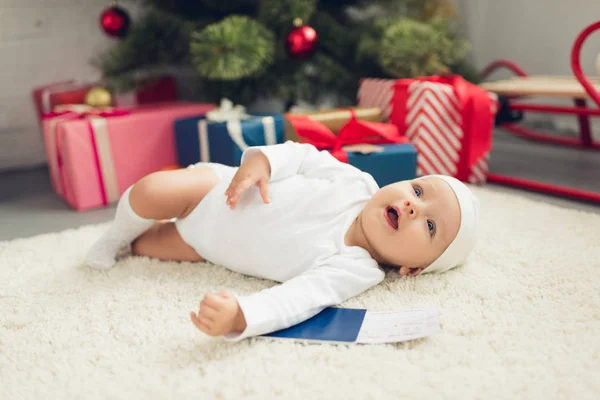Adorable little baby with flight ticket and passport lying on floor with christmas tree blurred on background — Stock Photo
