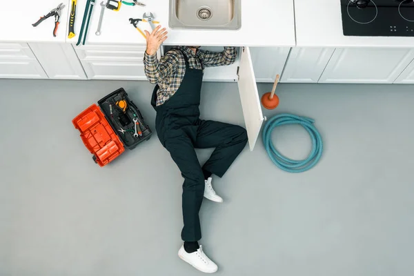 Vista ad alto angolo dell'idraulico sdraiato sul pavimento, controllando il lavandino e prendendo strumenti in cucina — Foto stock