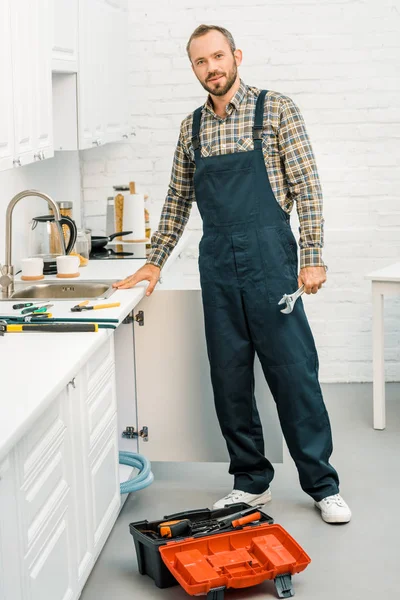 Beau plombier tenant clé réglable et regardant la caméra dans la cuisine — Photo de stock