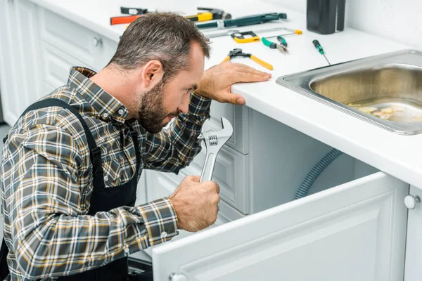 Barbudo reparador sosteniendo llave ajustable y mirando debajo del lavabo roto en la cocina - foto de stock