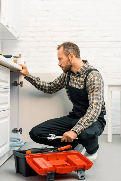 Seitenansicht des gutaussehenden Reparateurs mit verstellbarem Schraubenschlüssel und Blick unter kaputte Spüle in der Küche — Stockfoto