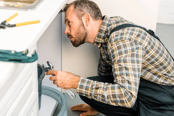 Vue latérale du beau plombier tenant une clé réglable et regardant sous l'évier cassé dans la cuisine — Photo de stock