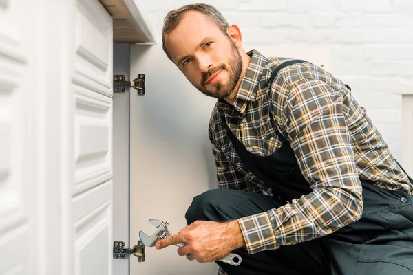 Beau plombier tenant clé réglable près de l'armoire de cuisine et regardant la caméra — Photo de stock