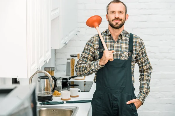 Fröhlich gut aussehender Klempner hält Kolben auf der Schulter und schaut in die Kamera in der Küche — Stockfoto