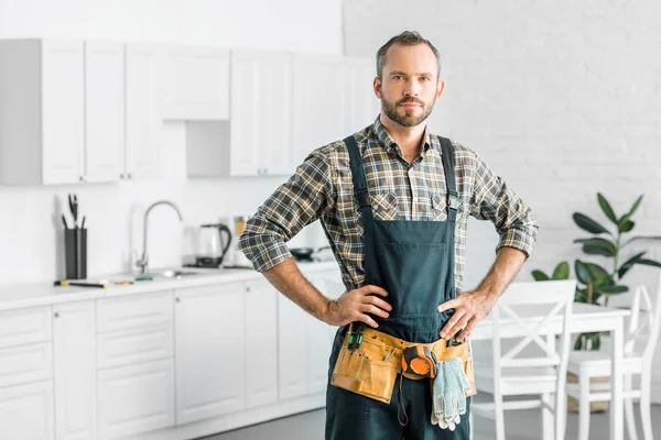 Schöner Klempner in Overalls und Werkzeuggürtel, der in der Küche in die Kamera schaut — Stockfoto