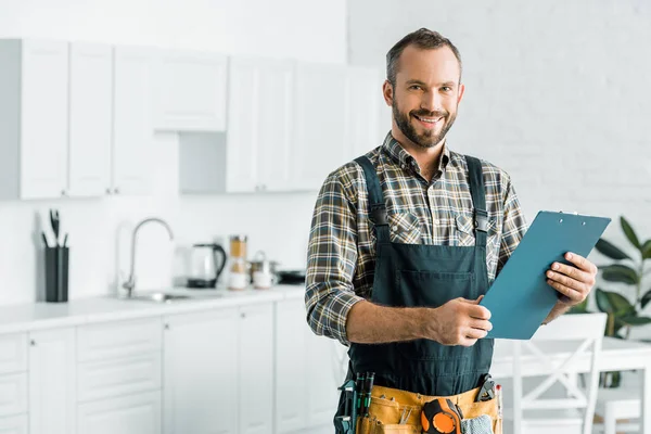 Lächelnd schöner Klempner mit Klemmbrett und Blick in die Kamera in der Küche — Stockfoto