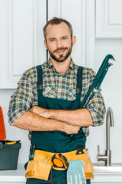 Fontanero guapo sosteniendo llave inglesa y mirando a la cámara en la cocina - foto de stock