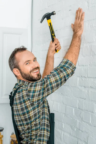 Sonriente reparador guapo clavo martillado en la pared blanca y mirando a la cámara - foto de stock