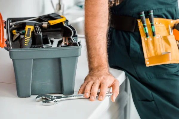 Abgeschnittenes Bild eines Klempners, der sich auf die Küchentheke lehnt und einen verstellbaren Schraubenschlüssel in der Küche berührt — Stockfoto